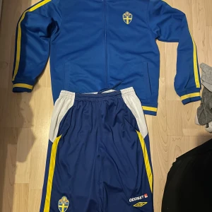 Blått träningsställ från Umbro - Säljer en blå träningsjacka från Umbro med Sveriges landslagsemblem på bröstet. Jackan har gula detaljer längs ärmarna och en dragkedja framtill. Perfekt för fotbollsträning eller som supporterplagg. Matchande shorts finns också tillgängliga. Säljes som ett set. Inga defekter, hör av er för mer info. 