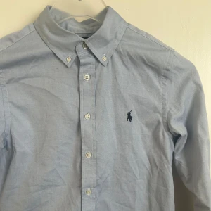 Blå skjorta från Ralph Lauren - Snygg ljusblå skjorta från Ralph Lauren med klassisk krage och knappar framtill. Skjortan har en broderad logotyp på bröstet och är långärmad. Perfekt för en stilren look. Perfekt nu till sommaren och den är i bra skick bara lite skrynklig.