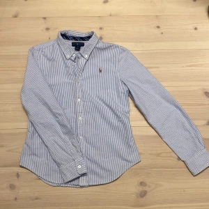  Raffe skjorta (Blåvit randig skjorta från Ralph Lauren) - Snygg blåvit randig skjorta från Ralph Lauren med klassisk krage och broderad logga på bröstet. Skjortan har långa ärmar och knappar framtill. Perfekt för en stilren look. I princip ny, inga defekter eller tecken på användning. Passar som XS.