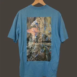 Acne Studios - Säljer en cool blå t-shirt från Acne Studios med ett unikt konstnärligt tryck på baksidan. T-shirten har en normal passform och är kortärmad, perfekt för en avslappnad stil. Den är gjord i mjukt bomullsmaterial och har en subtil logga på framsidan.