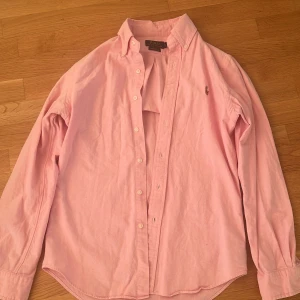 Rosa skjorta från Ralph Lauren - Säljer en stilren rosa skjorta från Ralph Lauren. Skjortan har en klassisk krage och knappar framtill. Den är broderad med den ikoniska loggan på bröstet. Perfekt för en elegant look.