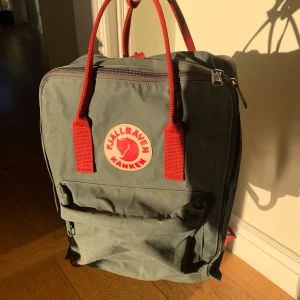 Grön ryggsäck från Fjällräven - En grön Fjällräven Kånken ryggsäck med röda handtag och axelremmar. Den har varit flitigt använd dock fortfarande i fint skick, nytvättad och alla dragkedjor samt spännen fungerar som de ska. Sittdynan som följer med original ligger kvar i väskan, den är dessvärre i sämre skick (se bild). Perfekt för vardagsbruk och utflykter. Skriv gärna privat vid frågor eller önskemål om fler bilder! 💕