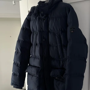 Mörkblå dunjacka från Tommy Hilfiger - Säljer en snygg mörkblå dunjacka från Tommy Hilfiger. Jackan har en hög krage och en huva för extra värme. Den är långärmad och har en diskret logga på ärmen. Perfekt för kyliga dagar!