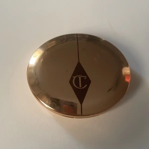 Puder från Charlotte Tilbury - Elegant puder i en lyxig guldskimrande dosa från Charlotte Tilbury. Nyansen är 'Fair' och dosan har en inbyggd spegel för enkel applicering. Perfekt för att sätta sminket och ge en jämn finish.