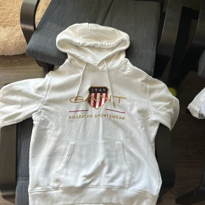 Vit hoodie från GANT - Säljer en vit hoodie från GANT med tryck på framsidan. Hoodien har en klassisk känguruficka och justerbar huva med dragsko. Perfekt för en avslappnad stil.