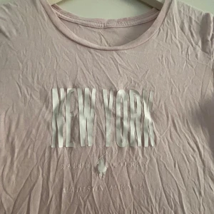 Ljusrosa t-shirt från Ralph Lauren - Säljer en ljusrosa t-shirt från Ralph Lauren med texten 'New York' tryckt på framsidan. T-shirten har en rund halsringning och korta ärmar. Perfekt för en avslappnad stil! Obs behövs strykas!