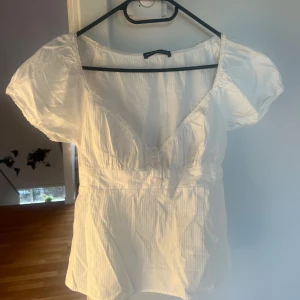 Vit blus från Brandy Melville - Säljer en söt vit blus från Brandy Melville med korta puffärmar och v-ringning. Blusen har en smickrande passform med lagom resår i midjan och en lätt rynkad effekt. Perfekt för en somrig look!