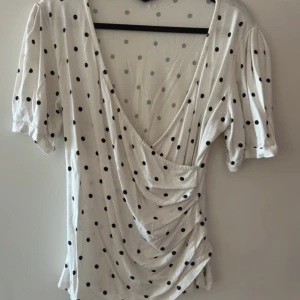 Vit prickig topp från Stockh LM - Säljer en vit topp med svarta prickar från Stockh LM. Toppen har en djup v-ringning och korta ärmar. Den är i en mjuk och bekväm bomullsmix, perfekt för en avslappnad stil.