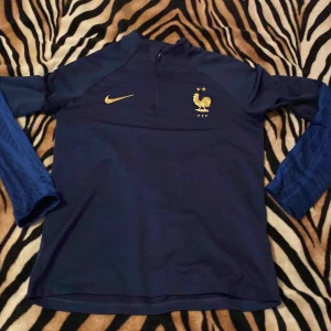 Blå fotbollströja och byxor från Nike - Säljer en blå fotbollströja och matchande byxor från Nike. Tröjan har långa ärmar och ett guldtryck av en tupp på bröstet. Byxorna har samma guldtryck och Nike-logga. Perfekt för fotbollsträning eller som supporterkläder. Tracksuiten är äkta och direkt från Nike köptes för 1 tusen. Säljer just nu för 749 men priset kan sänkas.