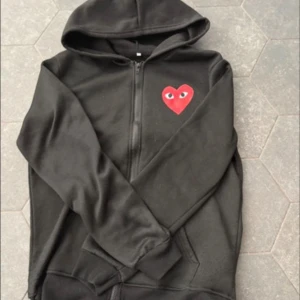 CDG hoodie - Säljer en svart hoodie med dragkedja och ett rött hjärta med ögon på bröstet. Perfekt för en avslappnad stil. Hoodien har långa ärmar och en bekväm passform.