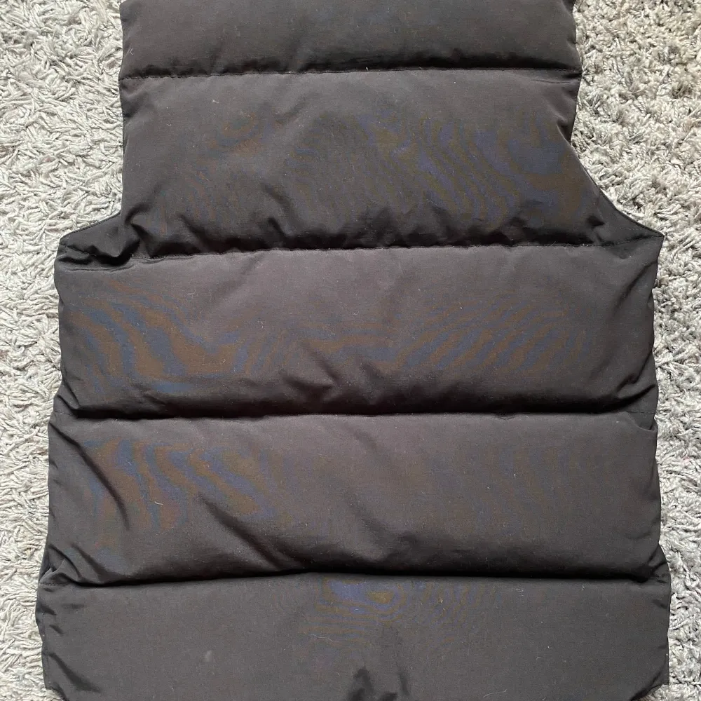 Säljer en svart dunväst från Canada Goose. Västen har en klassisk design med knappar och dragkedja framtill. Perfekt för kyligare dagar när du vill hålla värmen utan att kompromissa med stilen. . Takit.