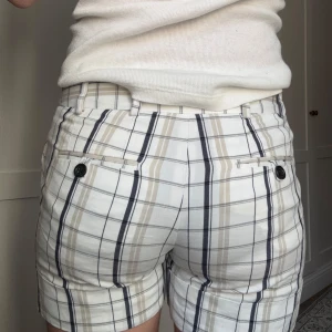 Rutig shorts från Zara Basic - Snygga rutiga shorts från Zara Basic i beige, svart och vit. De har en klassisk passform med knappdetaljer bak och är perfekta för en stilren look. Materialet känns bekvämt och passar bra för varmare dagar.