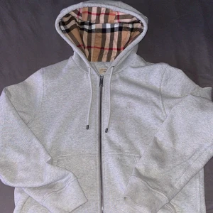 Grå hoodie från Burberry - Säljer en stilren grå hoodie från Burberry med klassiskt rutigt mönster i huvan. Tröjan har dragkedja och långa ärmar, perfekt för en avslappnad look. Passar bra till både jeans och joggers.