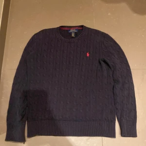 Mörkblå stickad tröja från Ralph Lauren - Säljer en snygg mörkblå stickad tröja från Ralph Lauren med kabelstickat mönster. Tröjan har långa ärmar och en rund halsringning. Den ikoniska röda loggan är broderad på bröstet. Perfekt för kyliga dagar och kall sommar kväller, priset går såklart även att diskutera och skriv gärna vid frågor 