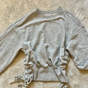 flätad tröja från Monki - Säljer en snygg grå tröja från Monki med coola snörningsdetaljer på sidorna. Tröjan har en normal passform och är tillverkad i ett mjukt material.