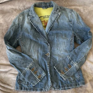 Figursydd jeansjacka - Snygg figursydd jeansjacka! med klassisk kavajdesign. Jackan har långa ärmar och knappar på ärmsluten samt framtill. Perfekt för en avslappnad men stilren look.