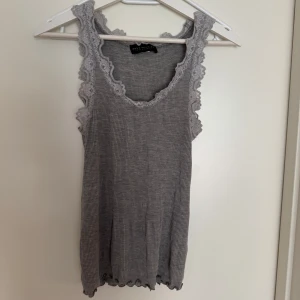 Grå ribbad linnetopp från Rosemunde - Säljer en stilren grå ribbad linnetopp från Rosemunde. Toppen har vackra spetsdetaljer längs ärmhålen och halsringningen, vilket ger en romantisk touch. Perfekt för lager-på-lager eller som den är under varma dagar.