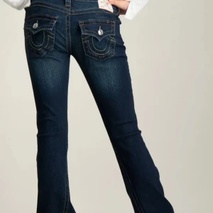 Mörkblå bootcut jeans, true religion jeans - Snygga mörkblå bootcut jeans med dekorativa fickor och knappar bak. Jeansen har en klassisk femficksdesign och en knappgylf framtill. Perfekta för en stilren look. Använda en gång, prislapp finns kvar, köpta för 1300kr