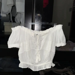 Vit offshoulder blus  - Säljer en söt vit offshoulder blus med korta ärmar och knytning framtill. Blusen har fina spetsdetaljer och en lätt volang vid ärmslut och nederkant. Perfekt för sommarens varma dagar! 🌸☀️🍹