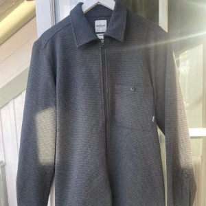 Grå overshirt från Dobber - Snygg grå overshirt från Dabber med dragkedja framtill och en bröstficka med knapp. Perfekt för lager-på-lager-stil. Den har en klassisk krage och långa ärmar.