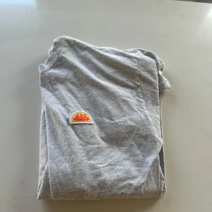 Grå t-shirt med broderad detalj - Säljer en grå t-shirt med en cool broderad detalj i form av en orange och gul sol på bröstet. Perfekt för en avslappnad stil. T-shirten har korta ärmar och är gjord i mjukt bomullsmaterial.