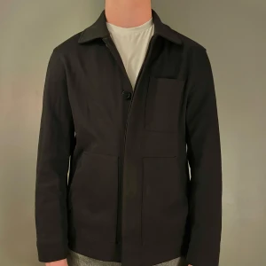 Svart overshirt från COS - Snygg svart overshirt från COS med en stilren design. Jackan har långa ärmar och knappar framtill. Perfekt för lager-på-lager-stil och passar till många olika outfits. Overshirten är i perfekt skick och passar perfekt inför sommaren. Modellen är 182 och väger cirka 76 kg! Hör av dig vid frågor!