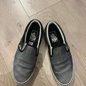 Glittriga slip-on sneakers från Vans - Snygga slip-on sneakers från Vans med en glittrig yta i blå och silver. Skorna har en klassisk vit sula och är perfekta för en avslappnad stil. De är lätta att ta på och av tack vare den elastiska designen. Strl 39. Glittret är lite skavd på ena skon (var så när de köptes)