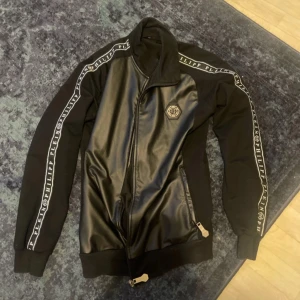 Svart jacka med läderdetaljer från Philipp Plein - Snygg svart jacka från Philipp Plein med läderdetaljer och dragkedja. Jackan har långa ärmar och logotyp längs ärmarna. Perfekt för en stilren look.