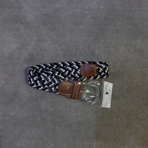 Svartvitt flätat bälte - Snyggt flätat bälte i svart och vitt med bruna läderdetaljer. Perfekt för att ge en stilren touch till din outfit. Bältet har ett klassiskt spänne i silverfärg.