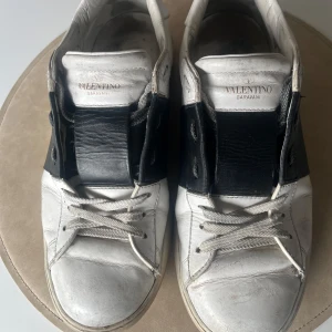 Valentino Garavani Opens - Säljer ett par stilrena Valentino Garavani sneakers i vitt och svart. Skorna har en bred svart rem över snörningen och klassiska nitar på hälen. Perfekta för en trendig look.