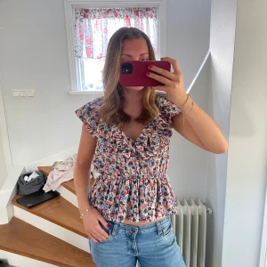 Blommig blus med volangdetaljer - Säljer en söt blommig blus med volangdetaljer och elastisk midja. Blusen har korta ärmar och ett färgglatt mönster i rosa, blått och vitt. Perfekt för en somrig look! 🌸