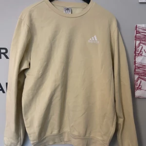 Beige/gul oversize sweatshirt från Adidas - Säljer en stilren beige sweatshirt från Adidas med rund hals och långärmad design. Perfekt för en avslappnad look. Adidas-loggan är tryckt i vitt på bröstet.
