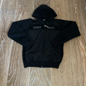 Svart huvtröja från Hugo Boss - Säljer en stilren svart huvtröja från Hugo Boss med dragkedja framtill. Tröjan har en klassisk design med logotypen på bröstet och en bekväm huva. Perfekt för en avslappnad look.