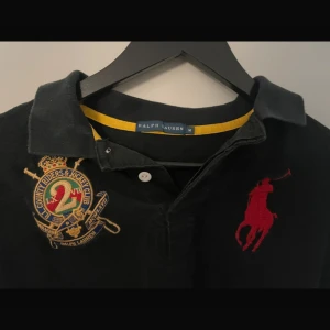 Svart pikétröja från Ralph Lauren - Säljer en svart pikétröja från Ralph Lauren med en stor röd logotyp på bröstet och ett broderat emblem på vänster sida. Tröjan har korta ärmar och en klassisk krage med knappar. Perfekt för en stilren look.