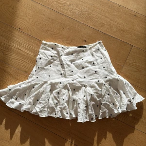 Vit kjol med volang och prickar - Charmig vit kjol med volangdetaljer och små svarta prickar. Perfekt för en somrig look med sin luftiga design och lekfulla mönster. Den har en fläck på sig sedan tidigare 