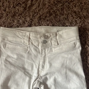 Vita bootcut jeans från H&M - Snygga vita bootcut jeans från H&M med justerbar midja och låg midja. Perfekta för en stilren look. De har en klassisk design med knappar och dragkedja framtill.