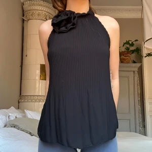 Svart topp med rosett💞 - Snygg svart ärmlös topp med ribbad struktur och en stor dekorativ rosett vid halsen. Perfekt för en stilren look.