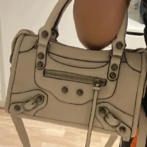 Beige handväska med dragkedja - Snygg beige/vit handväska med detaljerade sömmar och dragkedja framtill. Väskan har både handtag och en justerbar axelrem för flexibilitet. Perfekt för att bära dina dagliga nödvändigheter med stil.