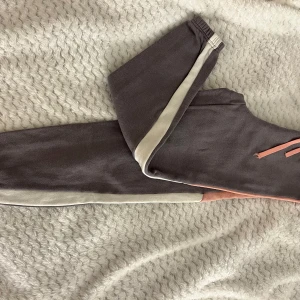 Grå mjukisbyxor från H&M - Säljer ett par grå mjukisbyxor från H&M med rosa snörning i midjan. Byxorna har en bekväm passform och är perfekta för en avslappnad stil. De har elastisk midja och ribbade muddar vid bensluten.