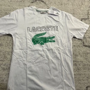 Vit t-shirt från Lacoste med grönt tryck - Säljer en vit t-shirt från Lacoste med det klassiska gröna krokodiltrycket och texten 'Lacoste' på framsidan. Perfekt för en stilren och sportig look.passar S 