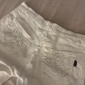 Vita jeansbyxor med strass från Para - Snygga vita jeansbyxor från Para med glittriga strassdetaljer på både fram- och bakfickor. Byxorna har klassisk femficksmodell, bälteshällor och dragkedja med knapp. Perfekta för dig som vill sticka ut lite extra med detaljer.
