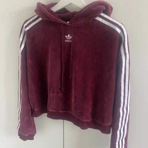 Croppad Adidas hoodie - Säljer en plommon lila hoodie från Adidas. Materialet är mjuk sammet med klassiska vita ränder längs ärmarna och broderad logga på bröstet. Tröjan har huva med snörning och är långärmad. 