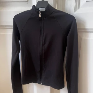 Svart zip-träningströja - Snygg svart tröja/kofta med dragkedja framtill. Den har en clean och minimalistisk look, perfekt för dig som gillar stilrena plagg. Långärmad och tillverkad i ett lätt och smidigt material som passar till många outfits. Endast använd 2-3 gånger och ser ut som ny!