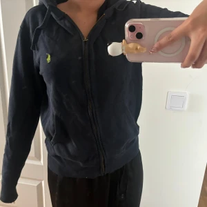 Mörkblå hoodiejacka från Ralph Lauren - Mörkblå hoodiejacka från Ralph Lauren med dragkedja framtill och klassisk huva. Jackan har en broderad grön logga på bröstet och kängurufickor. Perfekt för en chill och avslappnad stil. Passar som en S🩷