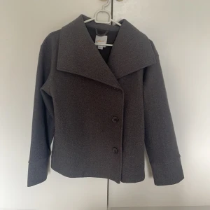 Grå kavajjacka från Gina Tricot - Snygg grå kavajjacka från Gina Tricot med bred krage och två knappar framtill. Jackan har en rak och lite boxig passform, långa ärmar och är gjord i ett mjukt ull-liknande material. Perfekt för dig som gillar stilrena och klassiska plagg.