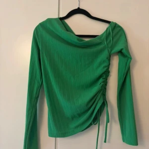 Grön ribbad topp från Gina Tricot - Snygg grön långärmad topp från Gina Tricot med ribbad struktur och trendig snörning på sidan. Toppen har en asymmetrisk halsringning och är croppad med en cool draperingseffekt. Perfekt för dig som vill sticka ut med färg och form!