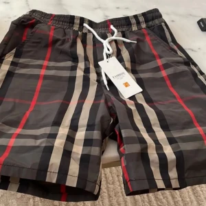 Burberry shorts  - Snygga shorts med klassiskt rutigt mönster i svart, beige och rött. De har elastisk midja med vit snörning för skön passform. Perfekta för sommaren och har en modern streetstyle-vibe. Materialet känns lätt och passar bra till varma dagar.