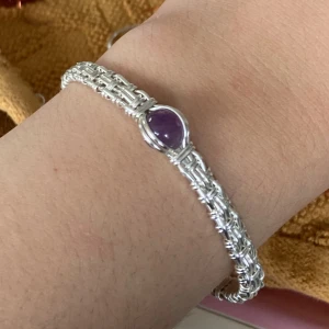 Handgjort silverarmband med lila sten - Unikt handgjort armband i flätad silverfärgad metalltråd med en oval, lila sten (ametist) i mitten. Armbandet har en snygg wire-wrapped design och är öppet i ändarna för enkel justering. Perfekt för dig som gillar boho eller statement-smycken.
