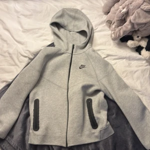 Grå hoodiejacka från Nike XS - Snygg grå hoodiejacka från Nike i storlek XS 158-170cm. Jackan har dragkedja framtill, två svarta fickor med dragkedja och en klassisk huva. Perfekt för chill dagar eller när du vill ha en sportig look. Materialet är mjukt och skönt, och Nike-loggan syns på bröstet.