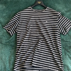 Randig t-shirt från J.Lindeberg - Svart och vit randig t-shirt från J.Lindeberg med broderad logga på bröstet. Klassisk passform med korta ärmar och rund hals. Tillverkad i mjuk bomull, perfekt för en clean och stilren look.